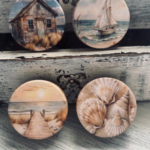 May include: Four round coasters with coastal designs. One features a weathered beach shack, another a sailboat on the ocean, a third a sunset over a boardwalk, and the last, a collection of seashells. The coasters are on a distressed white surface.