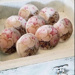 May include: Eight white ceramic drawer knobs with a floral design featuring pink roses and brown leaves. The knobs are arranged in a circle in a white wooden box.
