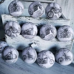 May include: A collection of round cabinet knobs with a vintage motorcycle design in black and white. The knobs feature a distressed, aged appearance, with the word "PATENTS" visible on some. The knobs are arranged on a distressed white wooden surface.