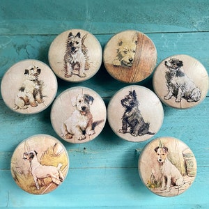 May include: Set of ten round wooden knobs with a variety of dog breeds printed on them. The knobs are painted in a light brown color and have a distressed finish. Each knob features a different dog breed, including a terrier, a dachshund, a pug, and a retriever.