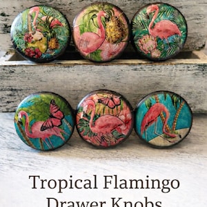 May include: Set of six round drawer knobs with tropical flamingo designs. Each knob features a pink flamingo with colorful tropical foliage, pineapples, and butterflies. The knobs are set against a white background with the text "Tropical Flamingo Drawer Knobs".