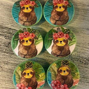 Op de afbeelding: Zes ronde houten knoppen voor lades met een groene achtergrond en een luiaard die een bloemenkrans draagt. De luiaards zijn bruin met gele gezichten en zijn omringd door groene bladeren.