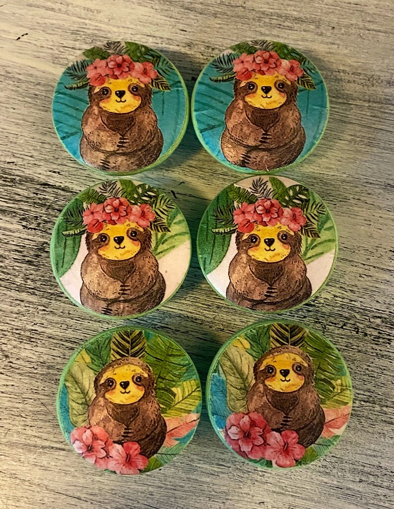 Op de afbeelding: Zes ronde houten knoppen voor lades met een groene achtergrond en een luiaard die een bloemenkrans draagt. De luiaards zijn bruin met gele gezichten en zijn omringd door groene bladeren.