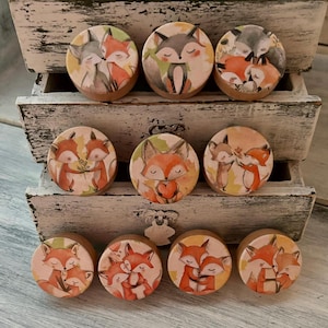 May include: Nine round wooden drawer knobs with fox illustrations. The knobs feature various scenes of foxes in different poses, including cuddling, holding hearts, and standing together. The knobs are painted in shades of orange, brown, and white.