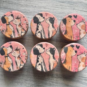 May include: Six round wooden cabinet knobs with dog and cat illustrations. Each knob features a different dog and cat pair, with a pink background and heart accents. The knobs are approximately 1.5 inches in diameter.