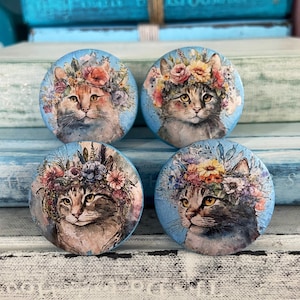 May include: Four round wooden ornaments with a blue background and a painted image of a cat wearing a flower crown. The cats are all different colors and have different expressions.