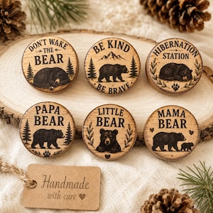 May include: Six circular wooden discs featuring burnt-in illustrations of bears and forest elements. Text includes: Don't Wake The Bear, Be Kind Be Brave, Hibernation Station, Papa Bear, Little Bear, and Mama Bear. A brown tag reads Handmade with care.
