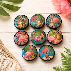 May include: Nine round decorative cabinet knobs with flamingo designs. Each knob features a different flamingo illustration with tropical plants and flowers in vibrant colors. The knobs are set against a white wooden background.
