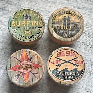 May include: Four wooden round decorative knobs with surfing-themed designs. Each knob features vintage-style graphics with text like "California Surfing Company" and "Big Sur Surf Team." The knobs have a weathered, rustic aesthetic.