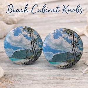 May include: Two round cabinet knobs with a beach scene design. The knobs feature a blue sky with clouds, palm trees, and a shoreline. The words "Beach Cabinet Knobs" are written at the top. Seashells and starfish surround the knobs.