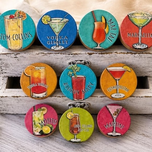 May include: A collection of nine round coasters featuring cocktail illustrations. Each coaster displays a different cocktail, including a Tom Collins, Margarita, and Mojito, with the cocktail's name printed on it. The coasters are arranged in a white wooden box.
