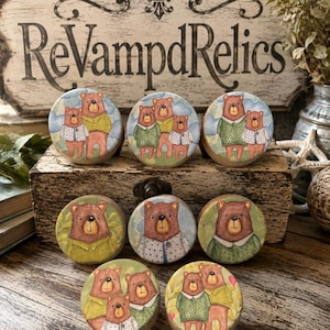 May include: A collection of eight round wooden drawer knobs featuring illustrations of brown bears in various clothing, including green sweaters and polka-dot shirts, set against soft-toned backgrounds of blue skies and green foliage.