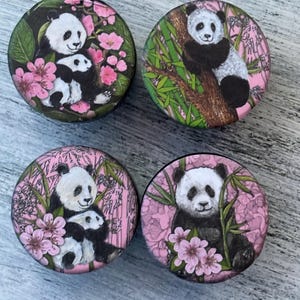 May include: Four round ceramic coasters with a pink background and a black and white panda bear design. Each coaster features a different panda bear pose, including a mother panda holding a baby panda, a panda sitting in a tree, and a panda sitting on the ground.