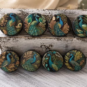 May include: Eight round decorative objects with peacock designs. Each features a peacock in shades of blue, green, and gold, set against a dark background with foliage. They are displayed in a distressed white wooden box.