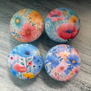 May include: Four round decorative knobs with floral designs. Each knob features a different watercolor floral pattern in shades of blue, pink, yellow, and orange. The knobs are set against a light blue background.