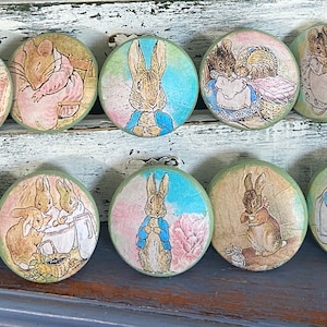 May include: Set of 10 round green wooden knobs with colorful illustrations of Peter Rabbit characters. The knobs feature Peter Rabbit, Benjamin Bunny, Mrs. Tiggy-Winkle, and other characters from the classic children's book.