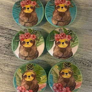 Op de afbeelding: Zes ronde, groene, keramische knoppen voor lades met een luiaard die een roze bloemenkroon draagt en op een groene bladerenachtergrond zit.