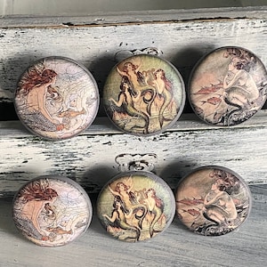 May include: Six round, gray, wooden drawer knobs with a mermaid design. Each knob features a different mermaid illustration with a vintage aesthetic.