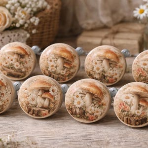 May include: Eight round cabinet knobs with a mushroom and floral design. The knobs have a cream background with a detailed illustration of mushrooms, daisies, and other flowers. The knobs are connected by a string of small, light blue beads.