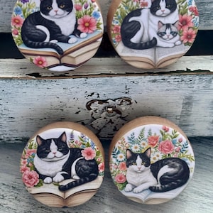 May include: Four wooden cabinet knobs with cat illustrations. Each knob features a different black and white cat design, surrounded by colorful flowers and resting on an open book. The knobs are round and have a natural wood finish.