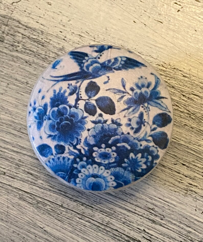 May include: A round, white ceramic knob with a blue and white floral design. The design features a bird, flowers, and leaves.