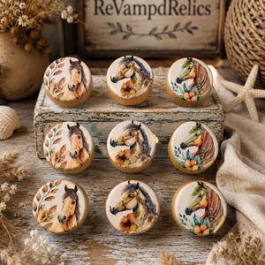 May include: Nine round wooden drawer knobs arranged in three rows of three. Each knob features a printed illustration of a horse head surrounded by botanical elements like leaves and flowers in warm earth tones and soft green accents.