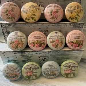 May include: Nine vintage-style round knobs with floral designs and text. The knobs are arranged in three rows on a wooden surface. The text on the knobs includes "Cafes & Legumes", "Maison", "Hotel Perrin", "Glaces Maison Clement", "Le Cacao Payraud", and "Mme Boileve".