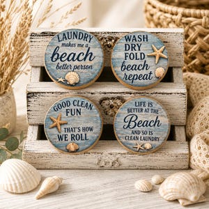 May include: Four round wooden signs with beach-themed quotes. Each sign has a blue and white wood grain background, a starfish, and a seashell. The quotes include "Laundry makes me a beach better person," and "Wash dry fold beach repeat."
