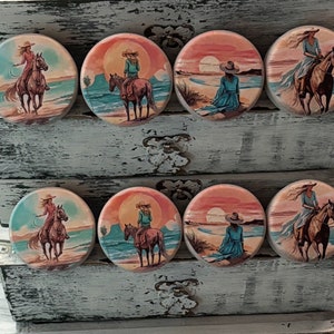 May include: Eight round wooden drawer pulls with a sunset scene and a woman on horseback. Each pull features a different color sunset and a woman in a different colored dress.