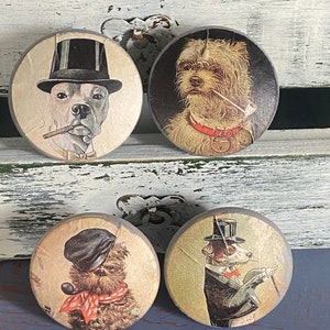 May include: Four round wooden knobs with different dog illustrations. The knobs feature a black and white dog wearing a top hat, a white dog with a red collar, a brown dog with a red scarf, and a black and white dog wearing a top hat and a bow tie.