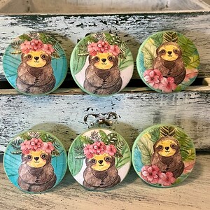 Op de afbeelding: Zes ronde houten knoppen met een groene achtergrond en een luiaard die een bloemenkrans draagt. De luiaards zijn bruin met gele ogen en zijn omringd door groene bladeren en roze bloemen.