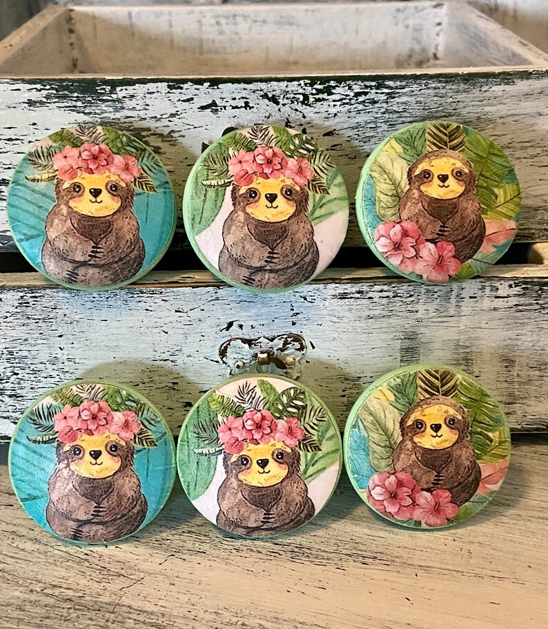 Op de afbeelding: Zes ronde houten knoppen met een groene achtergrond en een luiaard die een bloemenkrans draagt. De luiaards zijn bruin met gele ogen en zijn omringd door groene bladeren en roze bloemen.