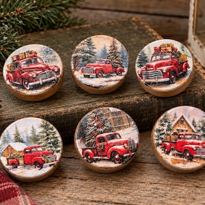 May include: A set of six round wooden knobs featuring printed illustrations of vintage red pickup trucks in snowy winter landscapes. Each knob displays a different scene with pine trees, holiday gifts, or small houses, all set against a snowy backdrop.