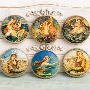 May include: Six round decorative cabinet knobs featuring vintage mermaid illustrations. Each knob showcases a different mermaid in a sea or ocean setting, with various colors and styles. The knobs are set against a white and light blue background.