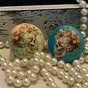 May include: Two decorative skull knobs with floral accents. One knob is on a light blue background and the other is on a cream background. Both knobs have a vintage look and are surrounded by white pearl beads.