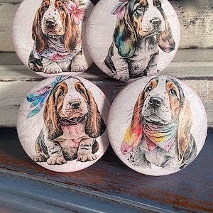 May include: Four round, white, ceramic knobs with a pink border. Each knob features a watercolor illustration of a basset hound puppy wearing a colorful bandana. The puppies are all in different poses.