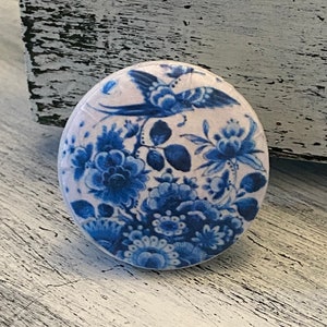 May include: A round ceramic knob with a white background and a blue and white floral design. The design features a bird in flight and a variety of flowers.