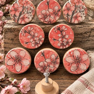May include: A set of eight round wooden drawer knobs featuring a coral-pink background with black-lined illustrations of cherry blossom flowers. Each knob has a flat circular face with a floral pattern and a cylindrical base with a metal screw.