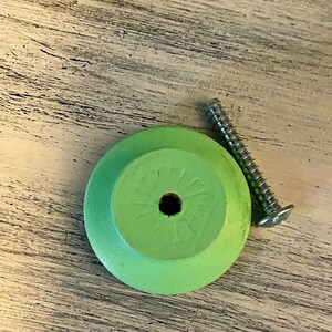 Op de afbeelding: Een ronde, lichtgroene houten knop met een schroefgat in het midden. De knop is bevestigd aan een zilveren schroef.
