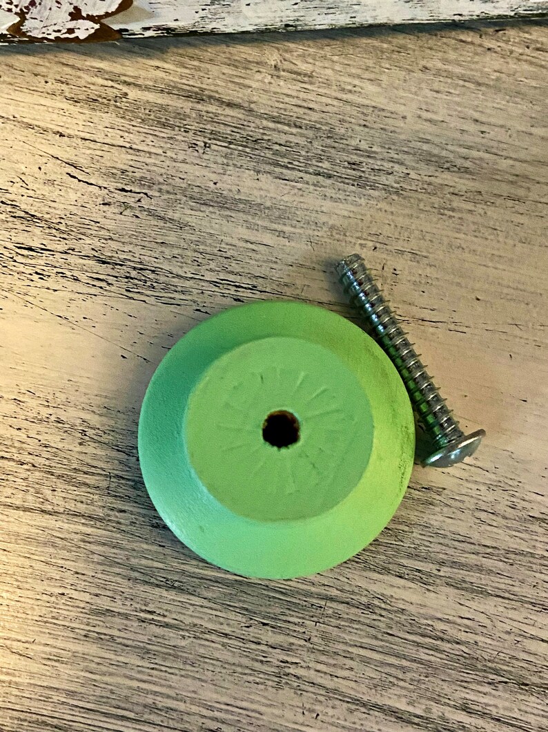 Op de afbeelding: Een ronde, lichtgroene houten knop met een schroefgat in het midden. De knop is bevestigd aan een zilveren schroef.
