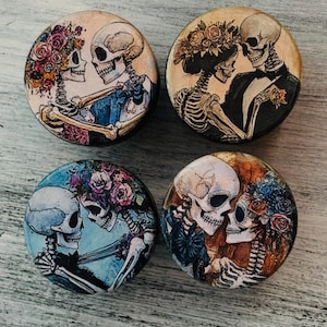 May include: Four round wooden ornaments with a painted design of skeletons dressed in wedding attire. The skeletons are holding hands and looking at each other. The ornaments are decorated with flowers and have a vintage aesthetic.