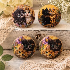 May include: Four circular wooden knobs featuring detailed illustrations of kittens. Each knob displays two kittens in various colors, including black, brown, and orange tabby, set against floral and golden backgrounds with intricate artistic patterns.
