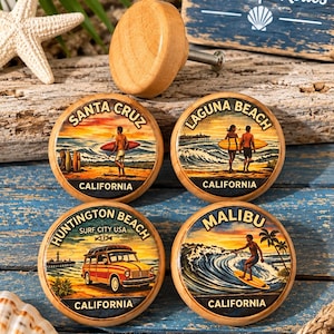 May include: Four round wooden cabinet knobs featuring colorful illustrations of California surf locations including Santa Cruz, Laguna Beach, Huntington Beach, and Malibu. Each knob displays a unique coastal scene with surfboards, waves, and vehicles.