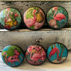 May include: Six round decorative cabinet knobs with tropical flamingo designs. Each knob features a different flamingo scene with pineapples, flowers, and butterflies. The knobs have a dark brown edge and are set against a white, distressed background.