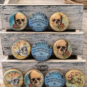 May include: A collection of round Ouija board planchette buttons with skull and floral designs. The buttons feature the text "OUIJA" and the alphabet. The color palette includes beige, blue, and red accents. The buttons are displayed in a rustic wooden box.