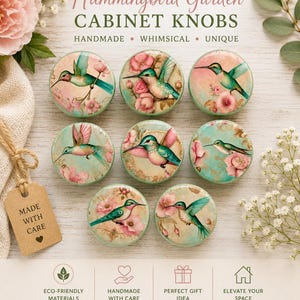 May include: A collection of eight round cabinet knobs featuring hummingbird designs with floral accents. The knobs have a light green border and are labeled "Hummingbird Garden CABINET KNOBS". The image also includes a tag that says "MADE WITH CARE".