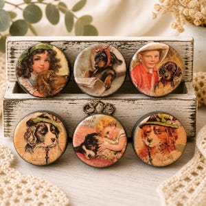 May include: A collection of six round vintage-style dog-themed decorative magnets. Each magnet features a different illustration of dogs and children, with a distressed, aged appearance. The magnets are displayed in a white wooden box.