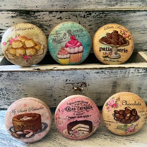 May include: Six round, decorative, wooden drawer pulls with a vintage, French-inspired design. Each pull features a different image of pastries, cakes, and chocolates. The pulls are painted in pastel colors with floral accents and text in French.