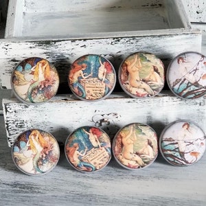 May include: Set of eight round drawer pulls with vintage mermaid illustrations. Each pull features a different mermaid design with a light blue, green, and brown color scheme. The pulls are made of wood and have a distressed finish.