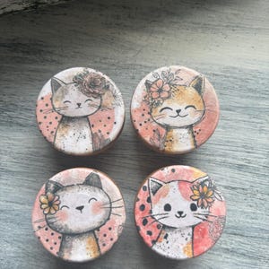 May include: Four round decorative cat designs. Each cat has a unique color scheme, with floral accents and a smiling face. The background is a soft pink with black polka dots. The designs are on a wooden surface.